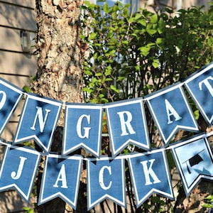 Op de afbeelding: Een blauwe en witte banner met de tekst "CONGRATS JACK" en afstudeerhoeden aan elk uiteinde.