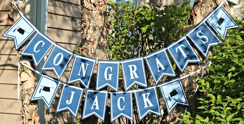 Op de afbeelding: Een blauwe en witte banner met de tekst "CONGRATS JACK" en afstudeerhoeden aan elk uiteinde.