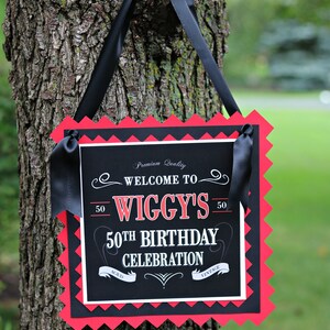 May include: Black and red sign with white text that says "Premium Quality Welcome to Wiggy's 50th Birthday Celebration Aged Vintage". The sign is hanging from a tree with black ribbon.