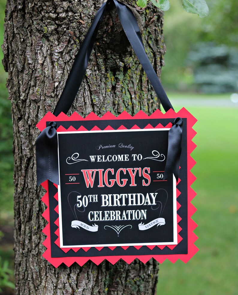 May include: Black and red sign with white text that says "Premium Quality Welcome to Wiggy's 50th Birthday Celebration Aged Vintage". The sign is hanging from a tree with black ribbon.