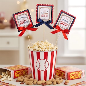 May include: A baseball-themed party setup with a red and white striped popcorn bucket, popcorn, and peanuts. Three decorative signs on sticks with text and bows are in the popcorn. Cracker Jack boxes are on the table.