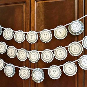 May include: A white and grey banner with the words "Happy 60th Anniversary Paul & 60" in a circle design. The banner is strung on a white ribbon and is hanging on a wooden door.