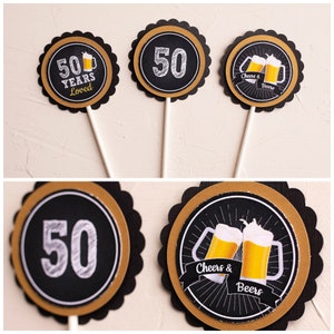Op de afbeelding: Set van drie zwarte, gouden en witte cupcake toppers. E&eacute;n zegt "50 Years Loved" met een bierpul, een andere heeft een witte "50" en de derde zegt "Cheers & Beers" met twee bierpullen. Elke topper zit op een witte stok.