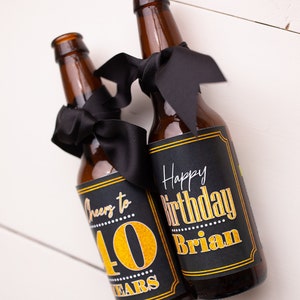 May include: Two brown glass beer bottles with black ribbon bows and personalized labels. The labels are black with gold lettering. One label says "Cheers to 40 Years" and the other says "Happy Birthday Brian".