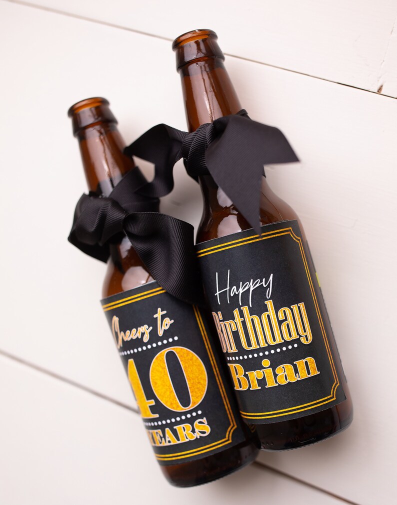 May include: Two brown glass beer bottles with black ribbon bows and personalized labels. The labels are black with gold lettering. One label says "Cheers to 40 Years" and the other says "Happy Birthday Brian".