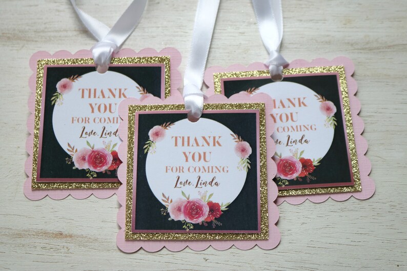 May include: Three pink and gold favor tags with a black background and floral design. The text on the tags reads "Thank You for Coming Love, Linda".