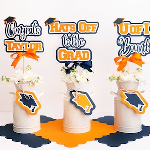 May include: Three graduation party centerpieces with white flowers in milk jugs. The centerpieces feature orange and navy blue graduation caps and text that reads "Congrats Taylor", "Hats Off to the Grad", and "U of I Bound".