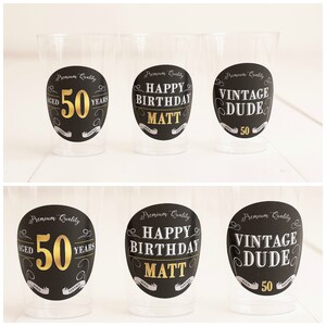 May include: Six clear plastic cups with black and gold circular labels. The labels have different text, including "Aged 50 Years", "Happy Birthday Matt", and "Vintage Dude".
