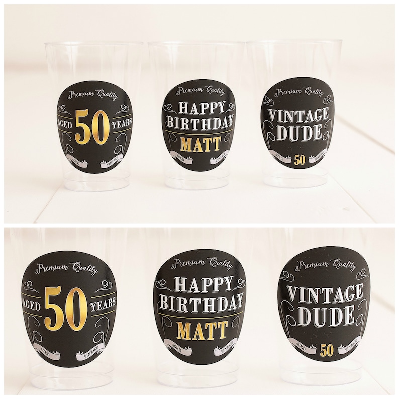 May include: Six clear plastic cups with black and gold circular labels. The labels have different text, including "Aged 50 Years", "Happy Birthday Matt", and "Vintage Dude".
