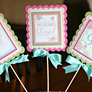 May include: Three decorative 90th birthday party centerpieces. Each has a scalloped edge in pink and green, with a square design in the center. The text reads "90 Years Loved", "90 Years Blessed", and "90". Each has a teal ribbon bow.