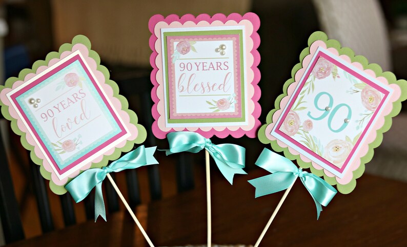 May include: Three decorative 90th birthday party centerpieces. Each has a scalloped edge in pink and green, with a square design in the center. The text reads "90 Years Loved", "90 Years Blessed", and "90". Each has a teal ribbon bow.