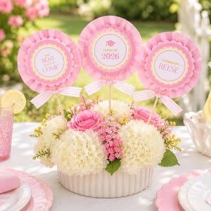 May include: A floral centerpiece with pink and white flowers in a white ceramic container. Three pink rosette decorations with text that reads "The Best is Yet to Come", "Class of 2026", and "Congrats Reese" are placed in the arrangement.
