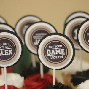 Puede incluir: Un conjunto de 10 adornos redondos para cupcakes con bordes negros y blancos y centros marrones. Los adornos presentan el texto "Feliz cumplea&ntilde;os" con un nombre, "Get Your Game Face On", y un tema deportivo.