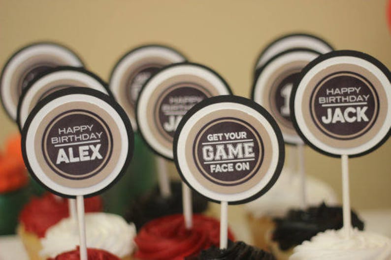 Puede incluir: Un conjunto de 10 adornos redondos para cupcakes con bordes negros y blancos y centros marrones. Los adornos presentan el texto "Feliz cumplea&ntilde;os" con un nombre, "Get Your Game Face On", y un tema deportivo.