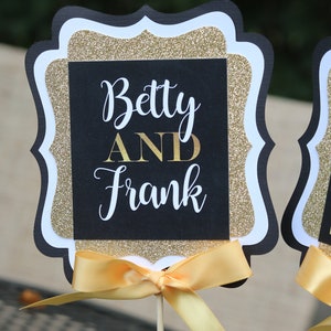 May include: Black and gold glitter table topper with a white border and a yellow ribbon. The topper says "Betty AND Frank" in white lettering.