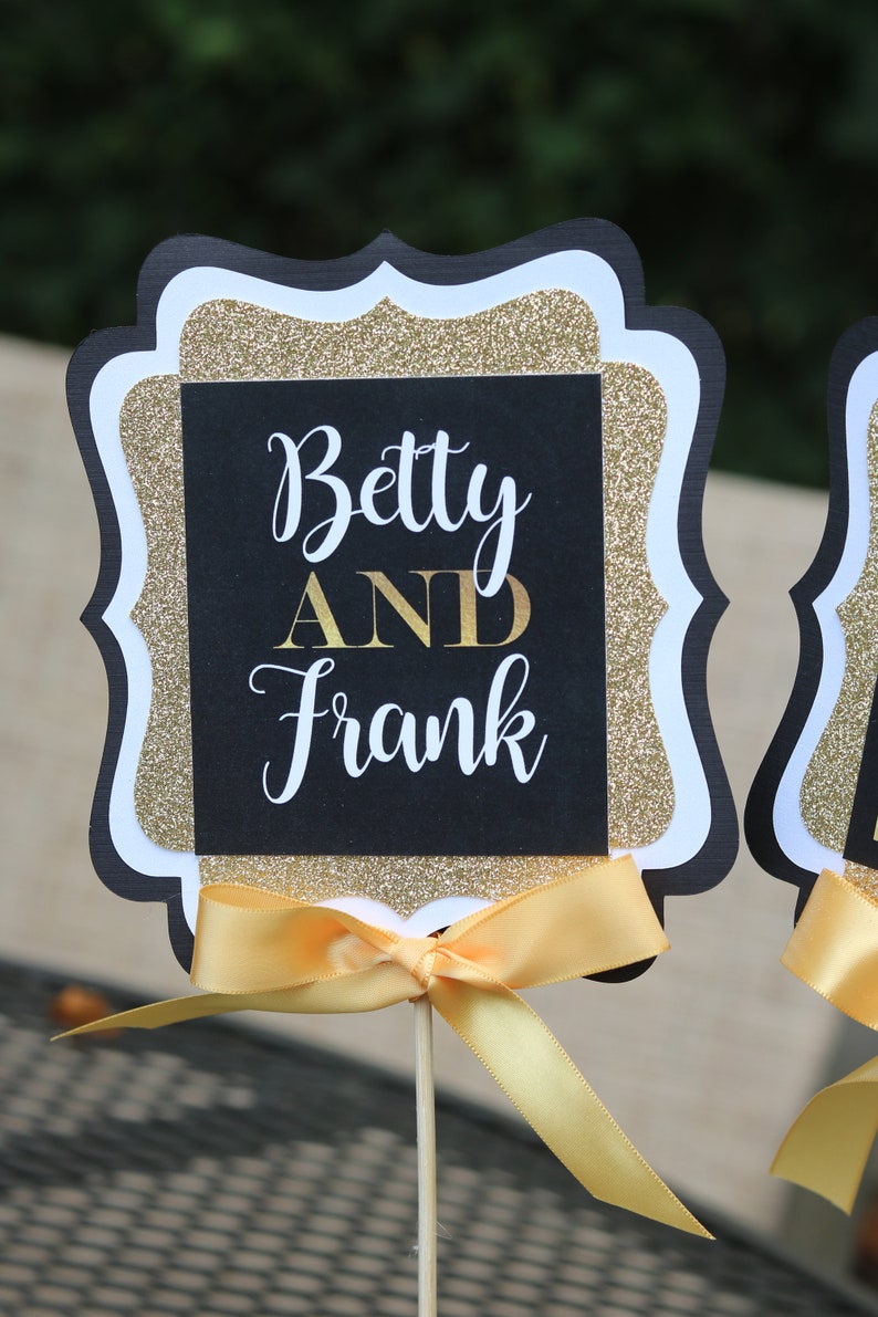 May include: Black and gold glitter table topper with a white border and a yellow ribbon. The topper says "Betty AND Frank" in white lettering.