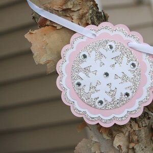 May include: A circular scalloped banner piece featuring a white snowflake cutout layered over a silver glitter circle and a light pink scalloped base. The snowflake is accented with small clear rhinestones and is strung on a white ribbon.
