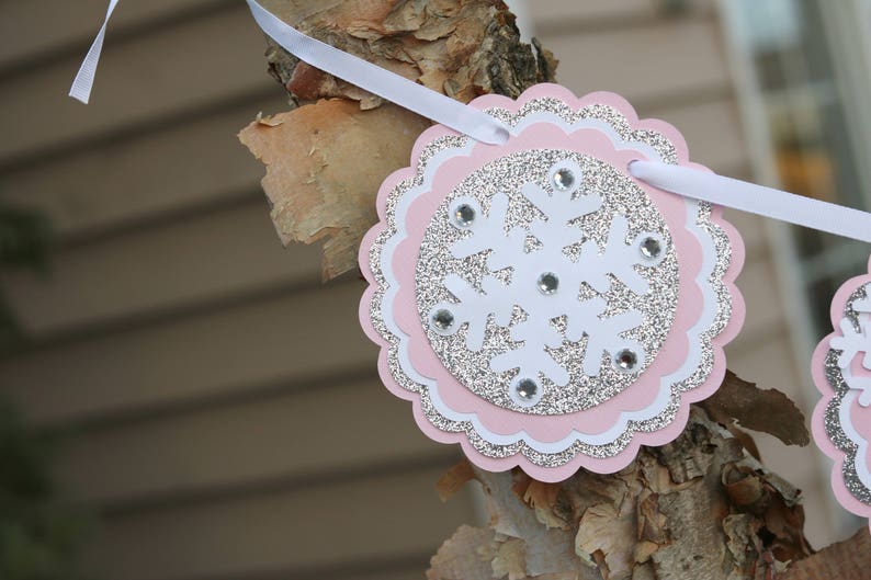 May include: A circular scalloped banner piece featuring a white snowflake cutout layered over a silver glitter circle and a light pink scalloped base. The snowflake is accented with small clear rhinestones and is strung on a white ribbon.