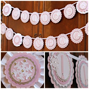 May include: A decorative banner featuring scalloped pink, white, and glittery gold paper circles strung on a white ribbon. The circles display individual letters spelling out "ELLIE'S ONE" with white snowflake cutouts and small clear rhinestones.