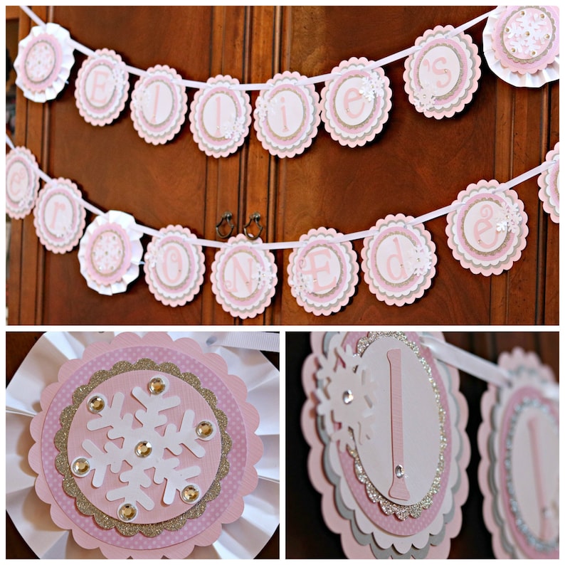 May include: A decorative banner featuring scalloped pink, white, and glittery gold paper circles strung on a white ribbon. The circles display individual letters spelling out "ELLIE'S ONE" with white snowflake cutouts and small clear rhinestones.