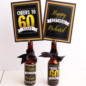 May include: Two black and gold signs with the text "Cheers to 60 Years" and "Happy Birthday Richard" on top of two brown glass bottles with black and white labels that say "Happy Birthday Richard" and "Cheers to 60 Years".