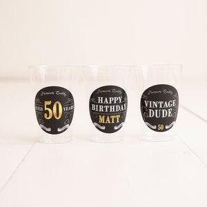 May include: Three clear plastic cups with black and gold labels. The labels say "Aged 50 Years", "Happy Birthday Matt", and "Vintage Dude 50".