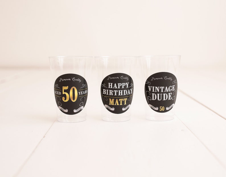 May include: Three clear plastic cups with black and gold labels. The labels say "Aged 50 Years", "Happy Birthday Matt", and "Vintage Dude 50".