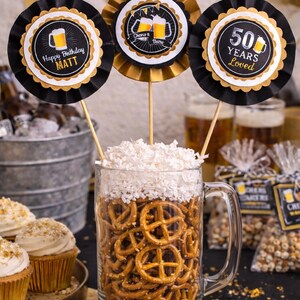 May include: A festive beer-themed party display. A large glass mug filled with pretzels and topped with a white foam-like substance. Three decorative toppers read "Happy Birthday Matt", "Cheers & Beers", and "50 Years Loved".