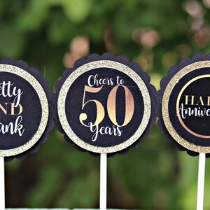 May include: Three black and gold cupcake toppers with scalloped edges. The first topper says "Betty AND Frank". The second topper says "Cheers to 50 Years". The third topper says "Happy Anniversary".