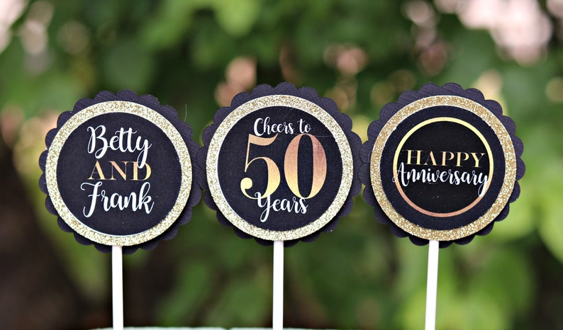 May include: Three black and gold cupcake toppers with scalloped edges. The first topper says "Betty AND Frank". The second topper says "Cheers to 50 Years". The third topper says "Happy Anniversary".