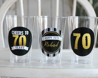 May include: Three clear plastic cups with black and gold labels. One label reads "Cheers to 70 Years", another says "Happy 70th Richard", and the third displays the number "70". The cups are for a 70th birthday celebration.