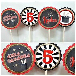 Op de afbeelding: Set van zes zwarte en rode cupcake toppers met gouden glitteraccenten. De toppers hebben een tovenaarshoed, sterren, het nummer 5 en de woorden "Abra Cadabra".