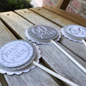 May include: Three silver and white cupcake toppers with a scalloped edge. One topper says "60 years", another says "Joan and Paul", and the third has a silver and white design with the number 60.