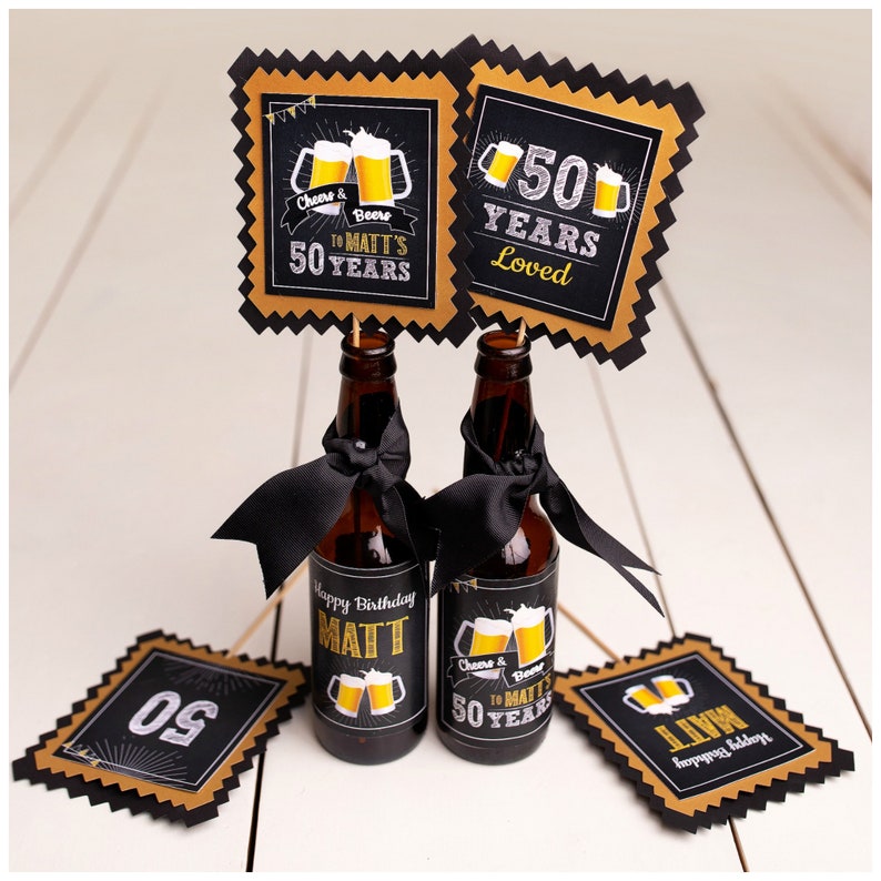 Op de afbeelding: Twee bruine bierflesjes met zwarte strikken en etiketten met de tekst "Happy Birthday MATT" en "Cheers & Beers to MATT'S 50 YEARS." Gouden en zwarte decoraties voor de 50e verjaardag.