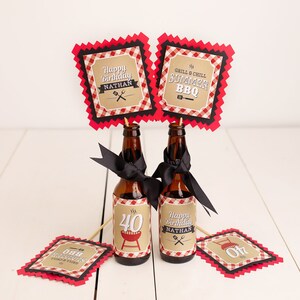 Peut inclure: Deux bouteilles de bi&egrave;re brunes d&eacute;cor&eacute;es de rubans noirs et d'&eacute;tiquettes personnalis&eacute;es. Les &eacute;tiquettes indiquent "Happy Birthday Nathan" et "Grill & Chill Summer BBQ". Des panneaux d&eacute;coratifs suppl&eacute;mentaires avec le m&ecirc;me texte sont sur des b&acirc;tonnets en bois. Le th&egrave;me est une f&ecirc;te d'anniversaire barbecue d'&eacute;t&eacute;.