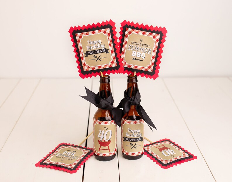 Peut inclure: Deux bouteilles de bi&egrave;re brunes d&eacute;cor&eacute;es de rubans noirs et d'&eacute;tiquettes personnalis&eacute;es. Les &eacute;tiquettes indiquent "Happy Birthday Nathan" et "Grill & Chill Summer BBQ". Des panneaux d&eacute;coratifs suppl&eacute;mentaires avec le m&ecirc;me texte sont sur des b&acirc;tonnets en bois. Le th&egrave;me est une f&ecirc;te d'anniversaire barbecue d'&eacute;t&eacute;.