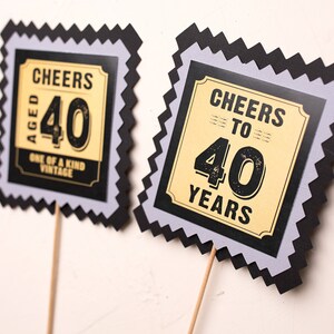 May include: Two black and grey scalloped cardstock signs with gold accents. The signs say "Cheers Aged 40 One of a Kind Vintage" and "Cheers to 40 Years".