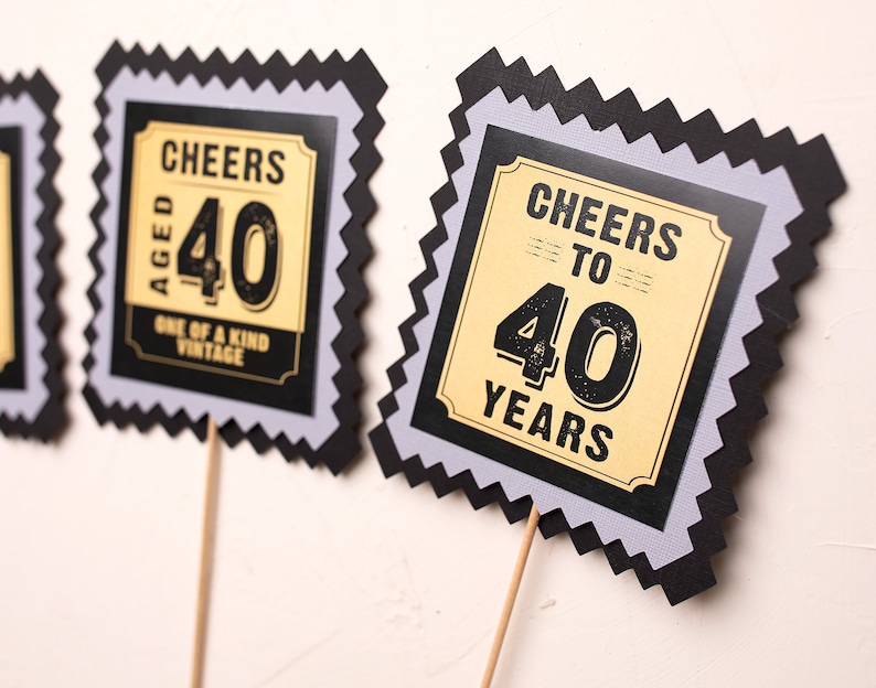 May include: Two black and grey scalloped cardstock signs with gold accents. The signs say "Cheers Aged 40 One of a Kind Vintage" and "Cheers to 40 Years".