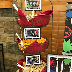May include: A three-tiered metal stand with red fabric liners holds three bowls of popcorn. Each bowl has a sign with a different flavor: Caramel Popcorn, Cheese Popcorn, and Butter Popcorn.
