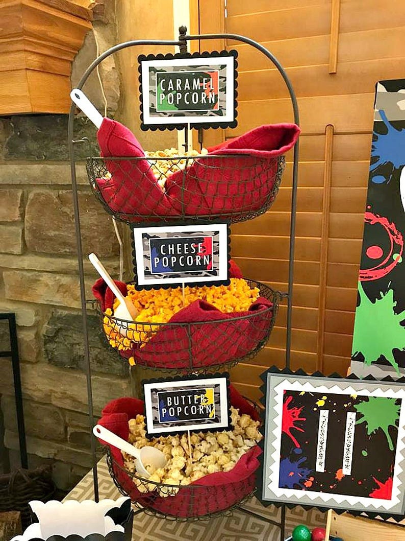 May include: A three-tiered metal stand with red fabric liners holds three bowls of popcorn. Each bowl has a sign with a different flavor: Caramel Popcorn, Cheese Popcorn, and Butter Popcorn.
