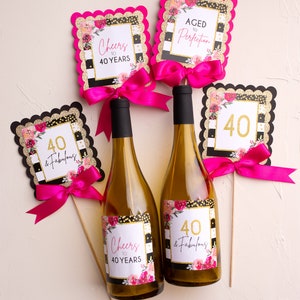 以下が含まれることがあります： 黒と白のストライプのラベルに金色のグリッターと花のアクセントが施されたワインボトルが2本。ラベルには「Cheers to 40 Years」と「40 & Fabulous」と書かれています。黒と白のストライプ、金色のグリッター、花のアクセントが施された装飾的なサインが4つあります。サインには「Cheers to 40 Years」、「Aged to Perfection」、「40 & Fabulous」、「40」と書かれています。