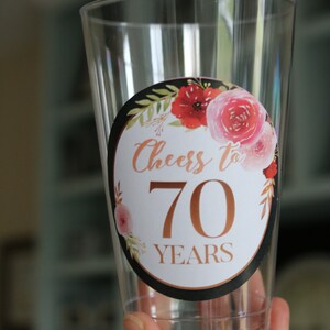 May include: Clear plastic cup with a round sticker featuring a floral design and the text "Cheers to 70 Years" in gold lettering.