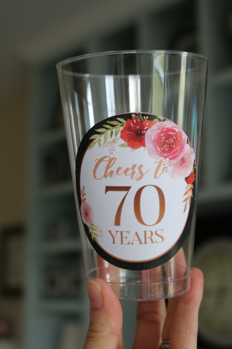 May include: Clear plastic cup with a round sticker featuring a floral design and the text "Cheers to 70 Years" in gold lettering.