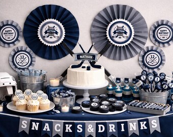 May include: A hockey-themed birthday party setup with a white cake, cupcakes, and various treats. Decorations include navy blue and gray paper fans, banners, and hockey pucks. The table is covered with a navy blue tablecloth, and the banner reads "SNACKS & DRINKS."