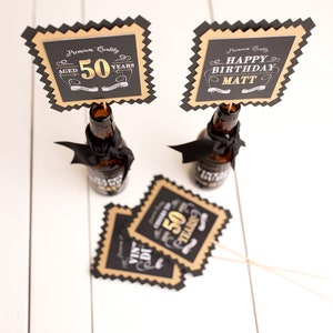 May include: Two black and gold party decorations with a bottle shape. One decoration says "Aged 50 Years" and the other says "Happy Birthday Matt".