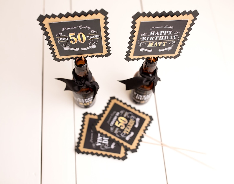 May include: Two black and gold party decorations with a bottle shape. One decoration says "Aged 50 Years" and the other says "Happy Birthday Matt".