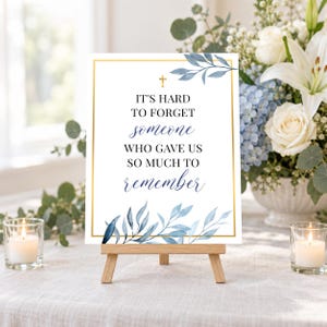 May include: A rectangular white sign with a gold border, featuring a small gold cross and blue watercolor leaves at the top and bottom. The text reads: IT'S HARD TO FORGET someone WHO GAVE US SO MUCH TO remember. The sign rests on a wooden easel.