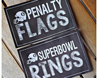 Può includere: Due cartelli neri con testo bianco e un'immagine di un casco da football. Il cartello superiore dice "Penalty Flags" e il cartello inferiore dice "Superbowl Rings".