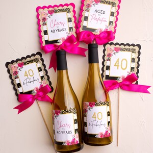 May include: Two wine bottles with labels that read "Cheers to 40 Years" and "40 & Fabulous." Decorative signs with the numbers "40" and phrases like "Aged to Perfection" are attached with pink bows. The signs have black, gold, and pink accents.