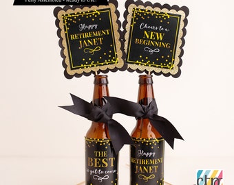 May include: Two black and gold retirement party decorations with the text "Happy Retirement Janet" and "Cheers to a New Beginning". The decorations are attached to beer bottles with black ribbon.